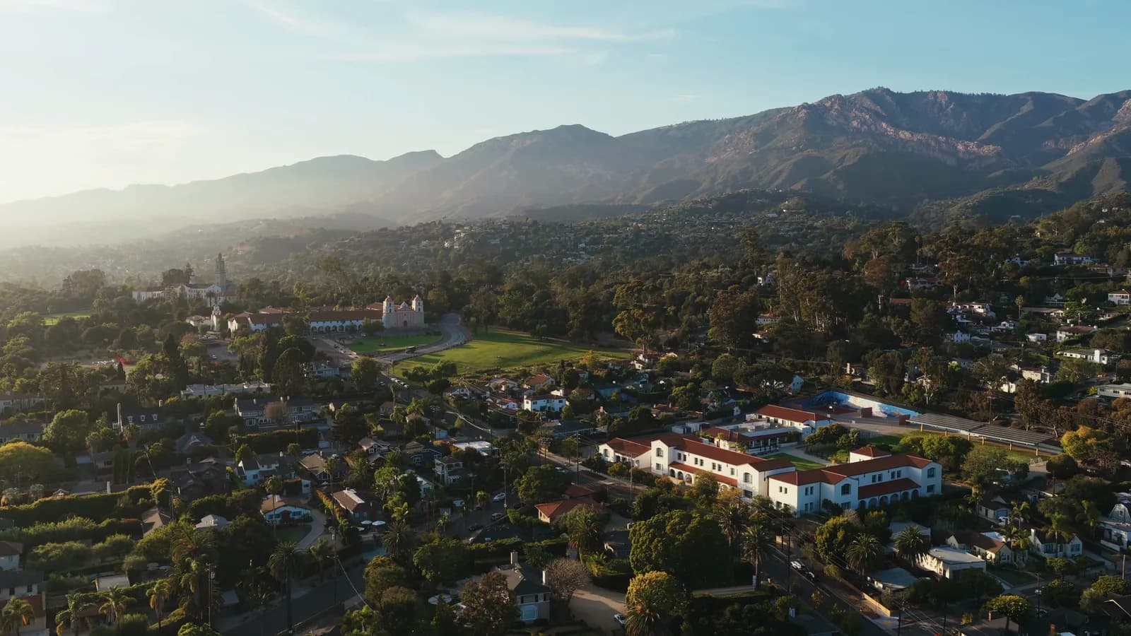 An aerial view over tree-lined neighborhoods and a mission, with mountains behind them in warm evening light.