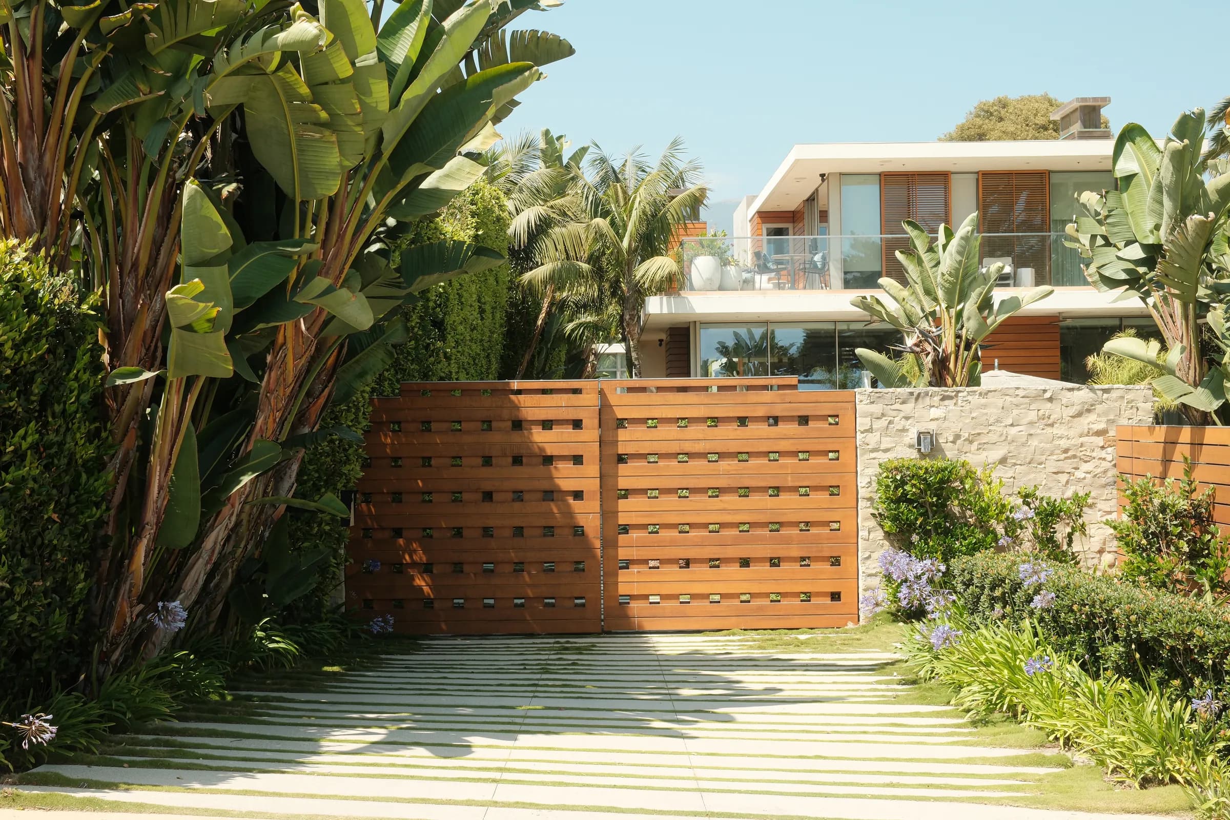 A contemporary house behind a black slatted fence with mature palms, and its interiors: a pale living room, an open kitchen and a detail of a patterned rug against leather. Frame 17 of 23.
